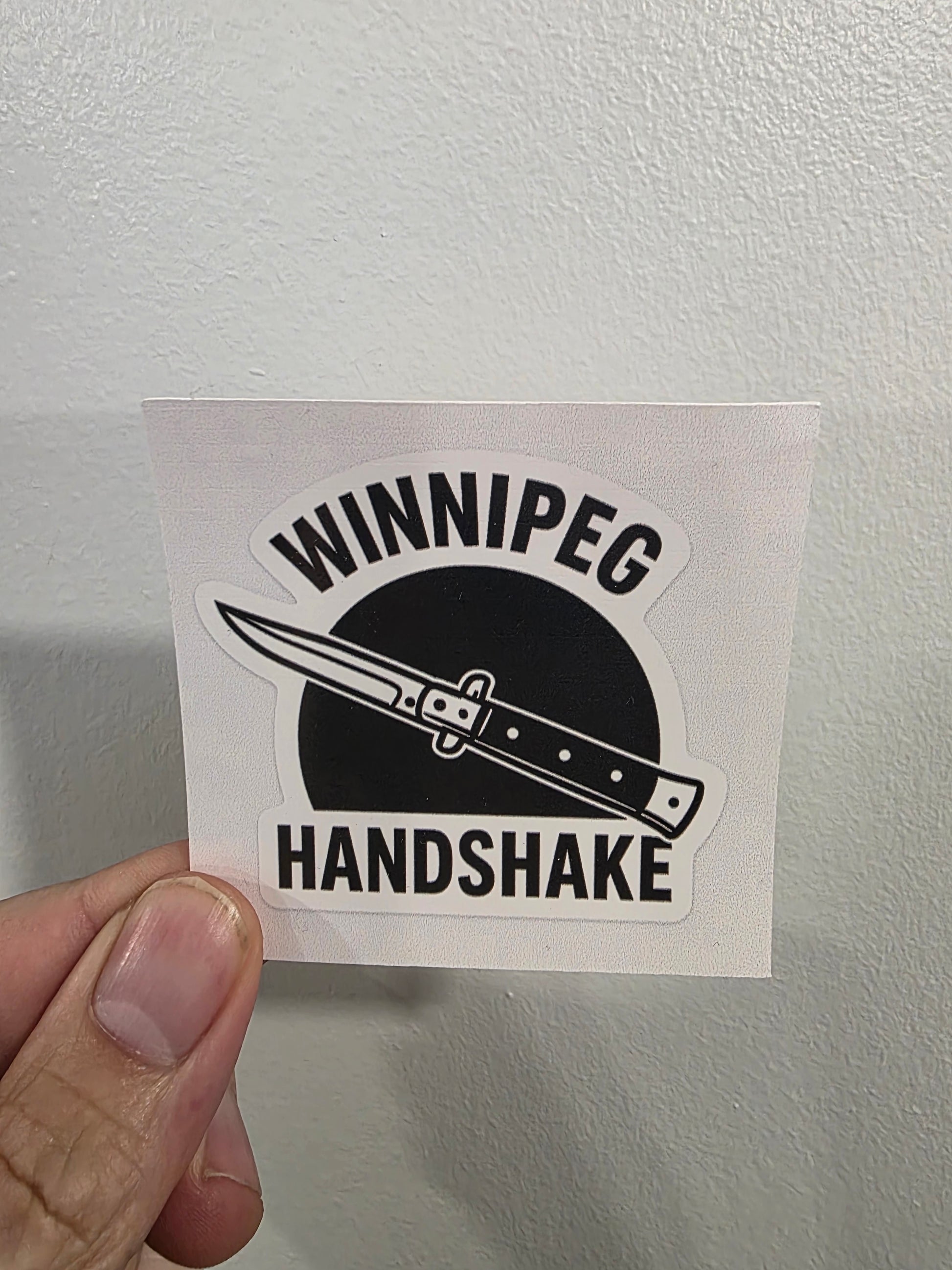 Sticker with a knife design and text held against a light gray wall.